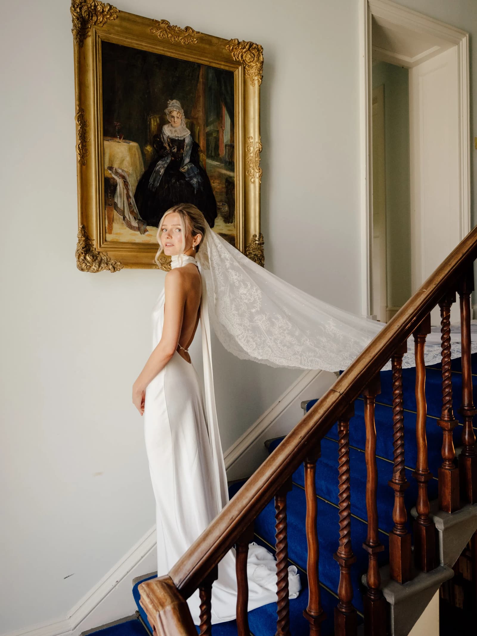 Bride on the staircase at Middleton House looking over her shoulder, her vintage silk veil draped across the banister