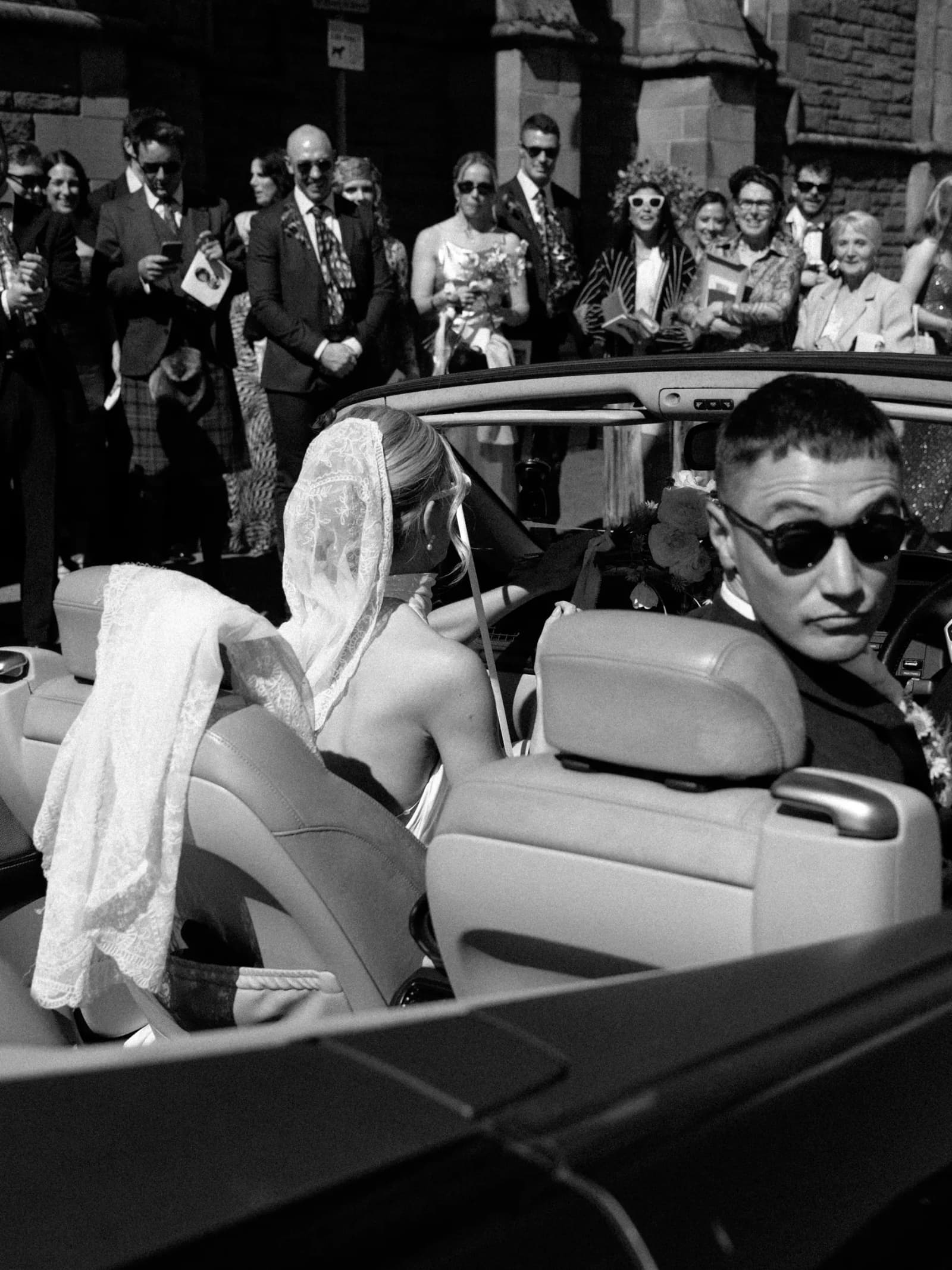 Groom in sunglasses at the wheel of an open-top car as the bride climbs in, cheered on by guests in Forfar, in black and white