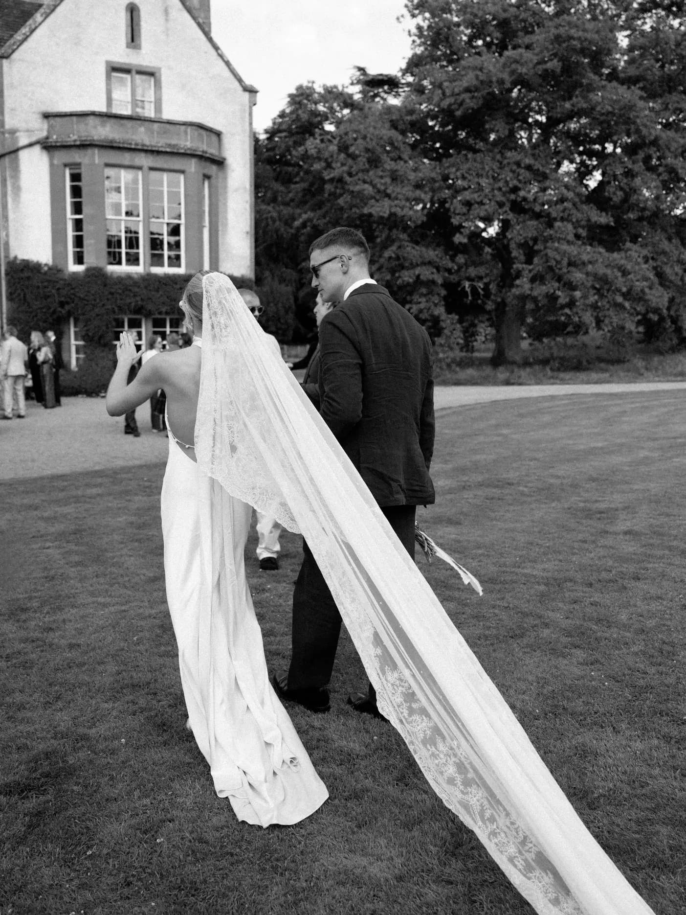 Bride and groom walking across the lawn at Middleton House, her cathedral-length veil trailing on the grass, in black and white