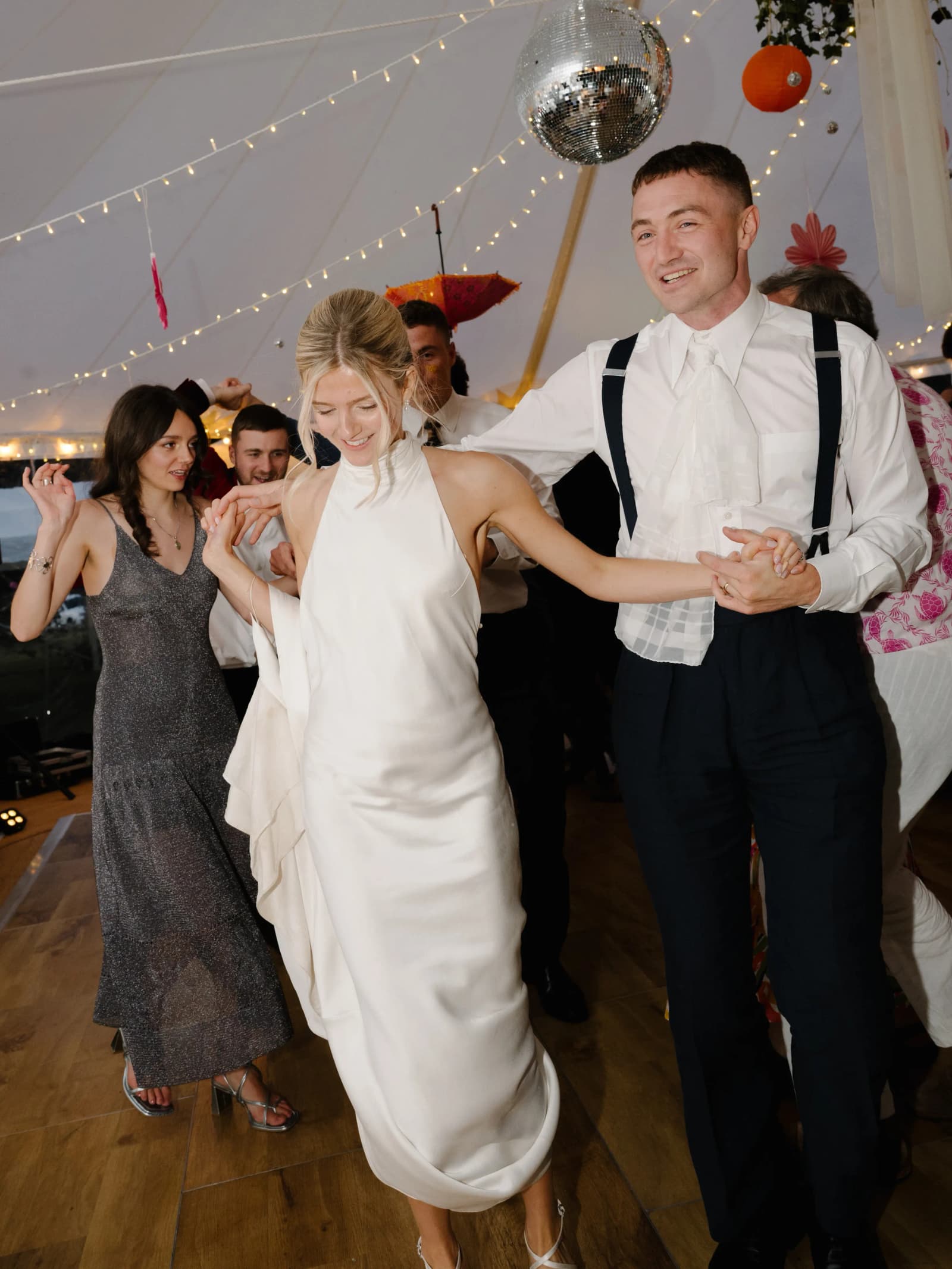 Bride and groom laughing and dancing with guests under festoon lights and a disco ball in a festival wedding marquee