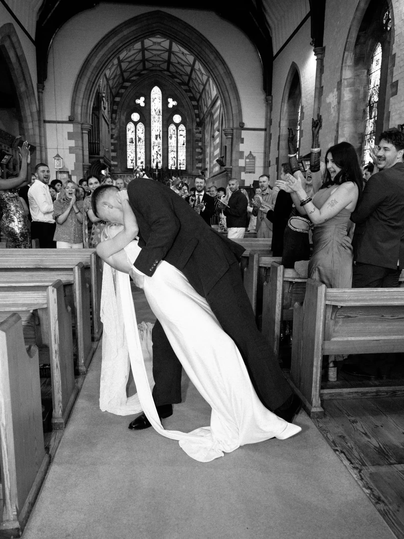 Groom dipping the bride for a kiss in the church aisle as guests cheer at a festival wedding in Scotland, in black and white