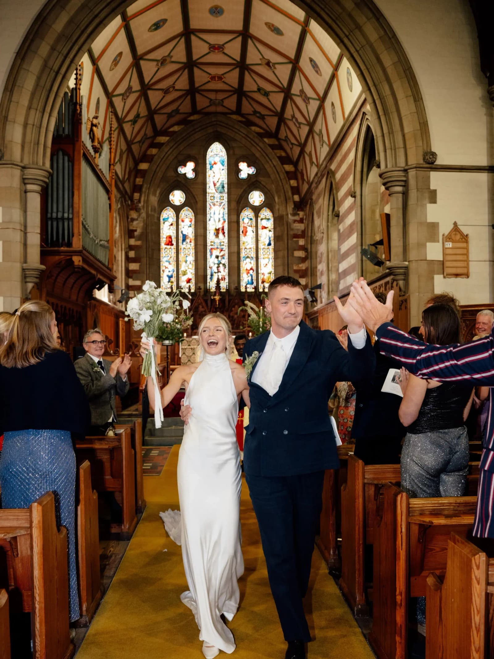 Newlyweds high-fiving guests as they walk back down the aisle beneath stained glass windows at their church wedding in Forfar