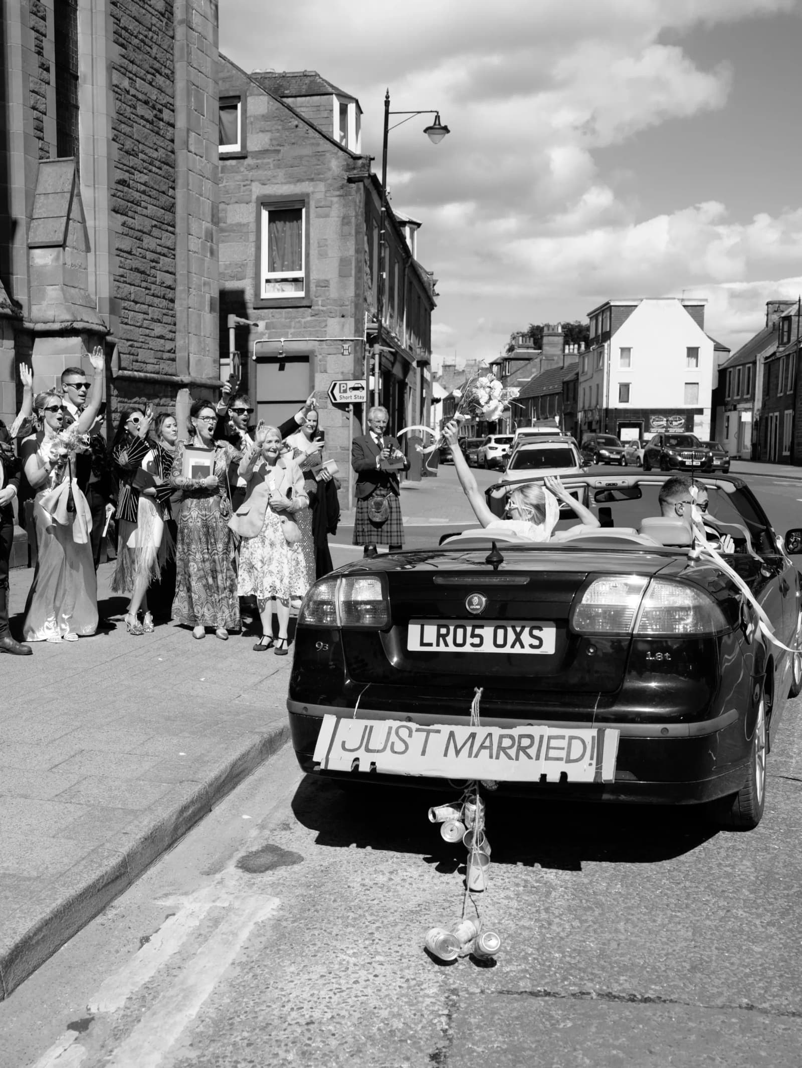Guests lining a street in Forfar to cheer the bride and groom off in their Just Married open-top car, in black and white