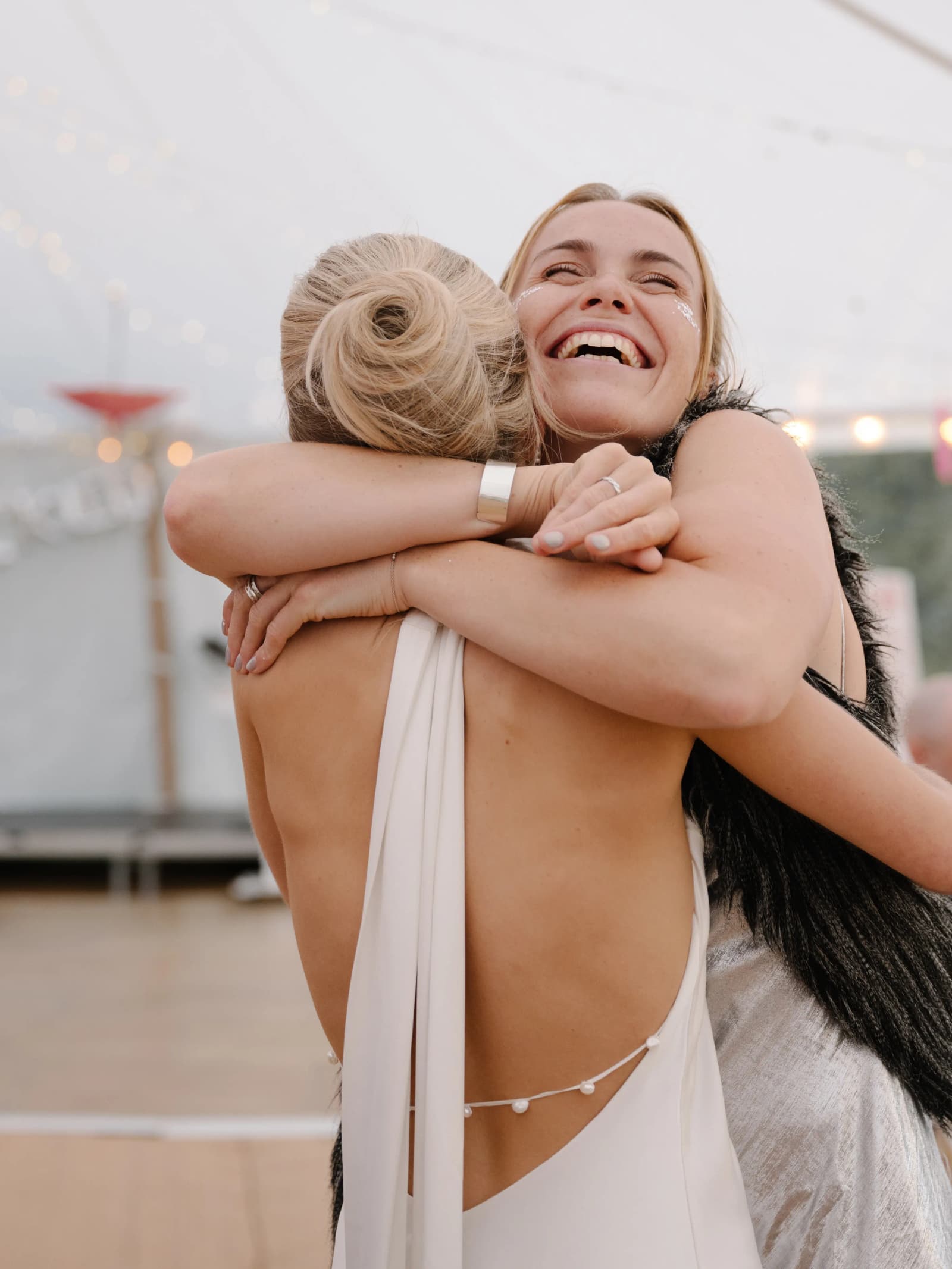 Bride in a backless gown sharing a big hug with a laughing guest in the marquee at a festival wedding in Scotland